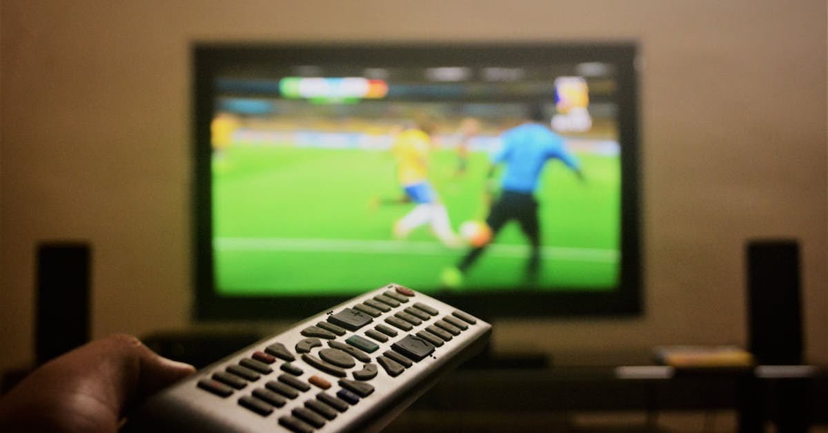 Close-up of a hand holding a remote control in front of a TV showing a football match.